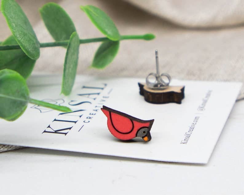 A small, red and black Cardinal Wood Stud Earring sits on a white Kinsal Creative card, with a green plant beside it. This handmade jewelry piece includes a matching earring back visible in the background.