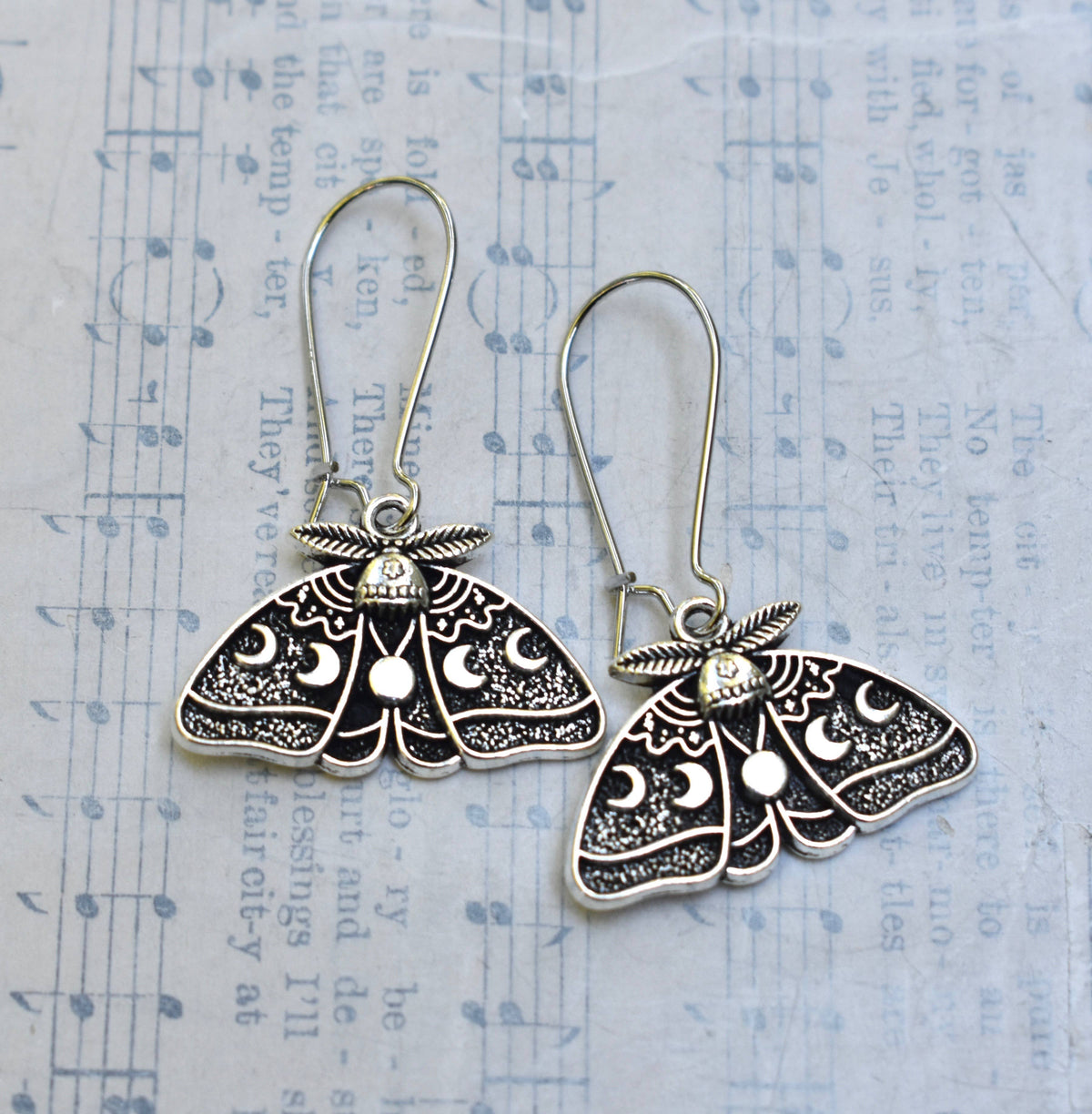 Luna Moth Earrings featuring delicate moon and star designs rest on sheet music, their mystical allure making them perfect goth statement earrings. Faded text and musical notes create a dreamy backdrop for these enchanting Luna Moth Earrings.