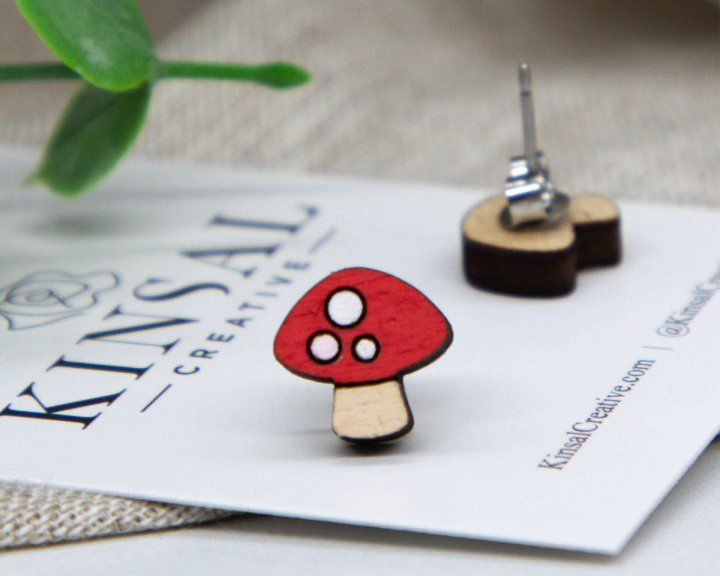 The Mushroom Wood Stud Earrings feature a red and white, mushroom-shaped design on wood, displayed on a KINSAL CREATIVE card. Another earring and green plant are in the background—ideal for those who love unique handmade jewelry.