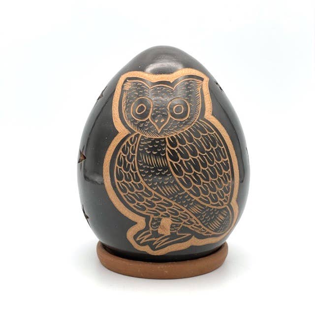 The Owl Luminary is a handcrafted, dark egg-shaped ceramic sculpture featuring a detailed owl engraving, displayed on a small brown base against a plain white background.