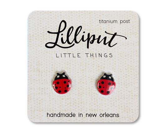 Ladybug Earrings: A pair of red ladybug-shaped stud earrings displayed on a card labeled “Lilliput Little Things,” featuring hypoallergenic titanium posts and “handmade in New Orleans” printed on the card. These charming Ladybug Earrings are gentle on sensitive ears thanks to their titanium posts.