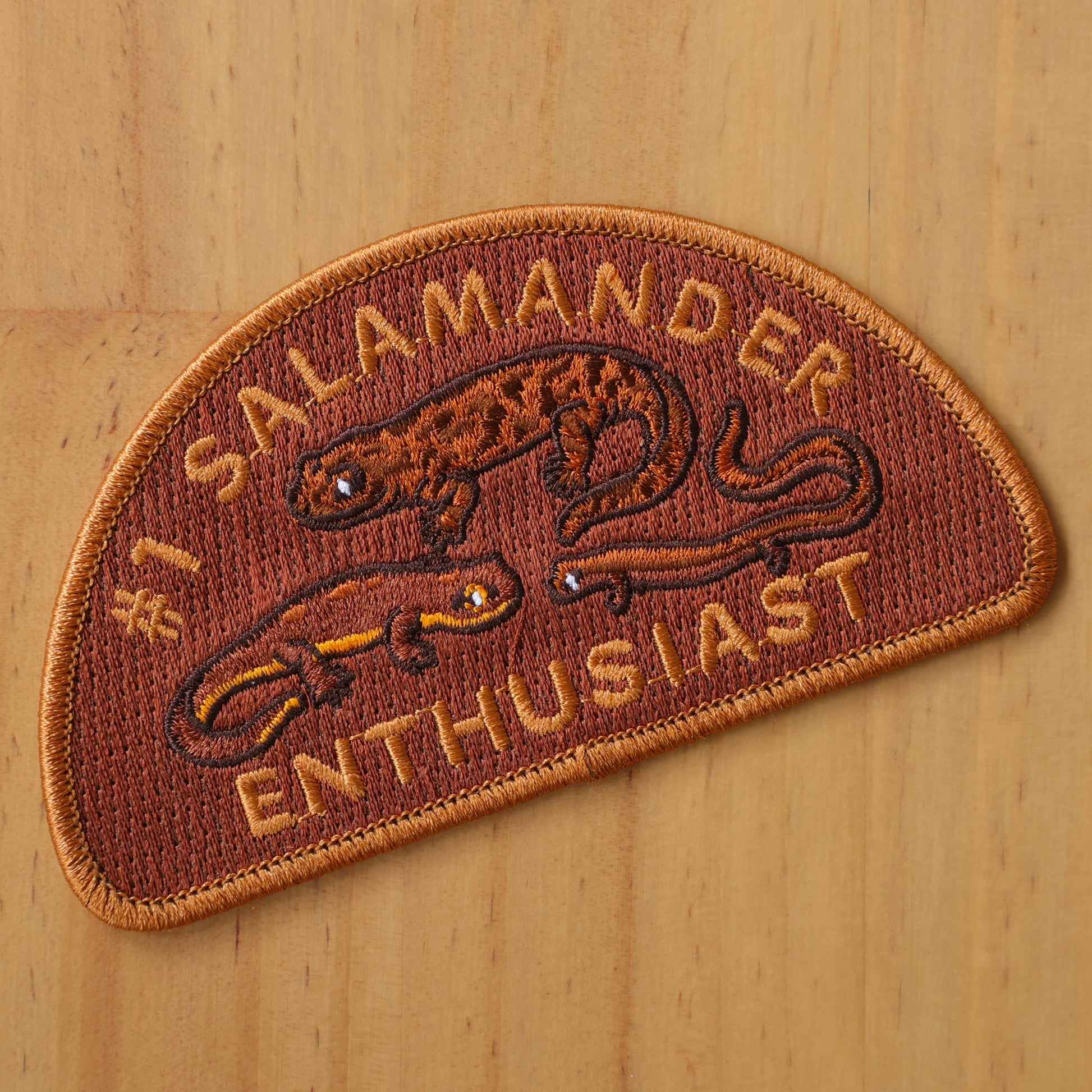 The #1 Salamander Enthusiast patch features two embroidered salamanders and yellow text on a reddish-brown background, shown on wood. Ideal as an iron-on patch for jackets or bags.