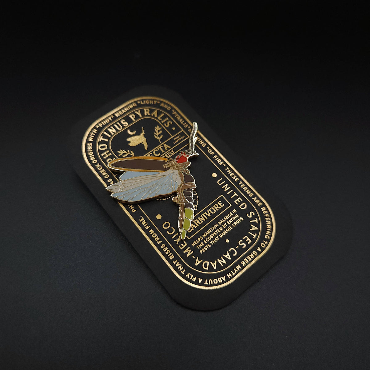 The Eastern Firefly Educational Insect Enamel Pin, featuring a gold and white moth design, is displayed on a black and gold card adorned with decorative text and a deer head motif, set against a dark background—making it a unique entomology gift.