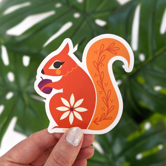 A hand holds a Vinyl Sticker Squirrel, an orange weatherproof die-cut sticker featuring a white flower on its hip and decorative leaf patterns on its tail.