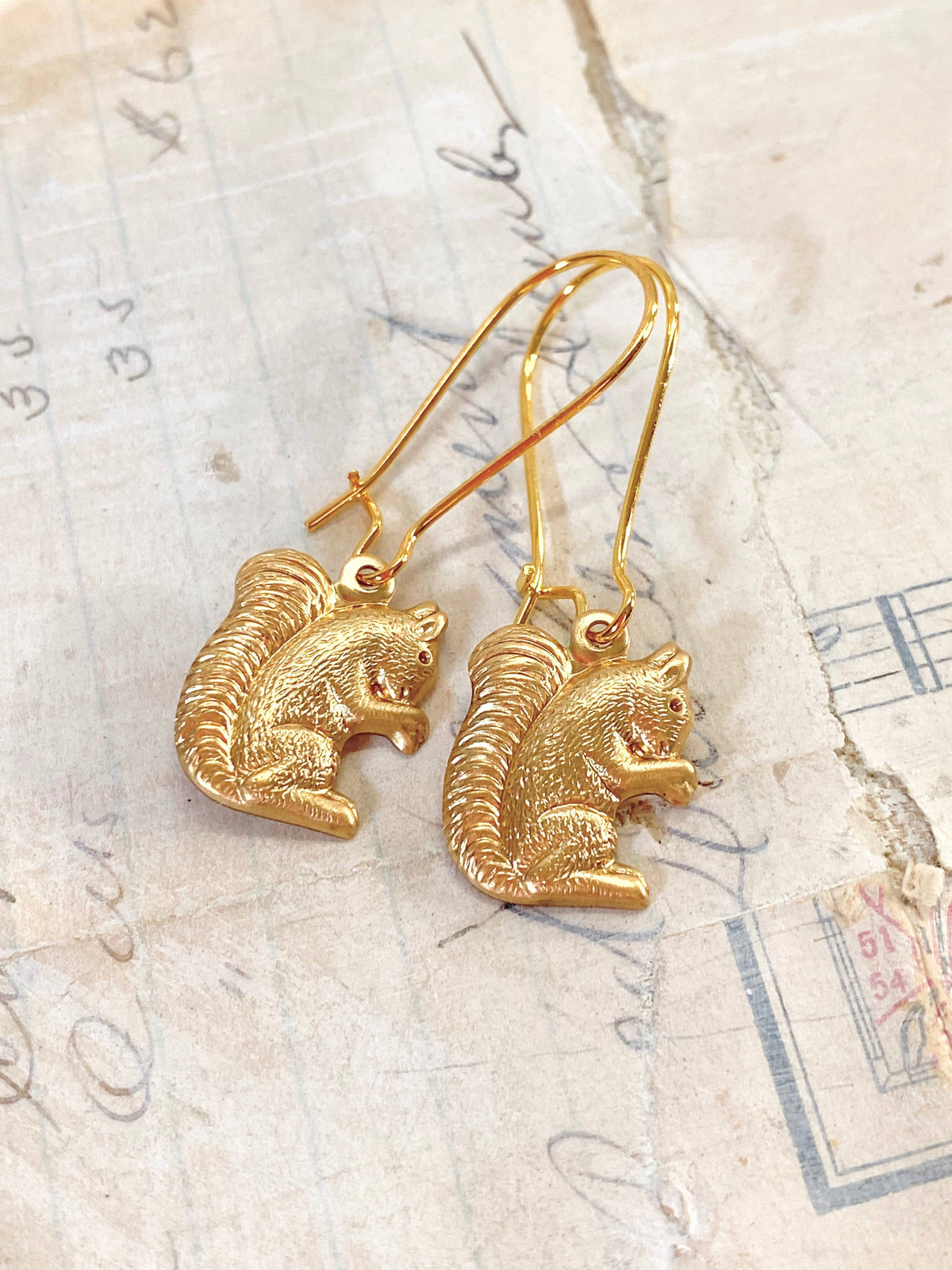 The Woodland Forest Squirrel Earrings rest on vintage paper adorned with handwritten notes and faded markings, capturing the whimsical charm of woodland jewelry inspired by forest creatures.