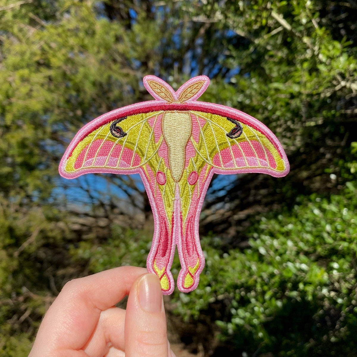 A hand holds a Pink Spirit Moth Iron-On Embroidered Patch with pink body and lime green wings against a blurred outdoor background.