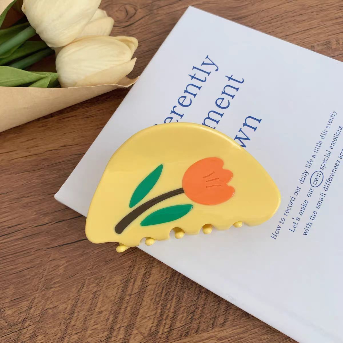 Yellow crescent-shaped Tulip Hair Claw Clip decorated with an orange flower and green leaves, resting on a white book.