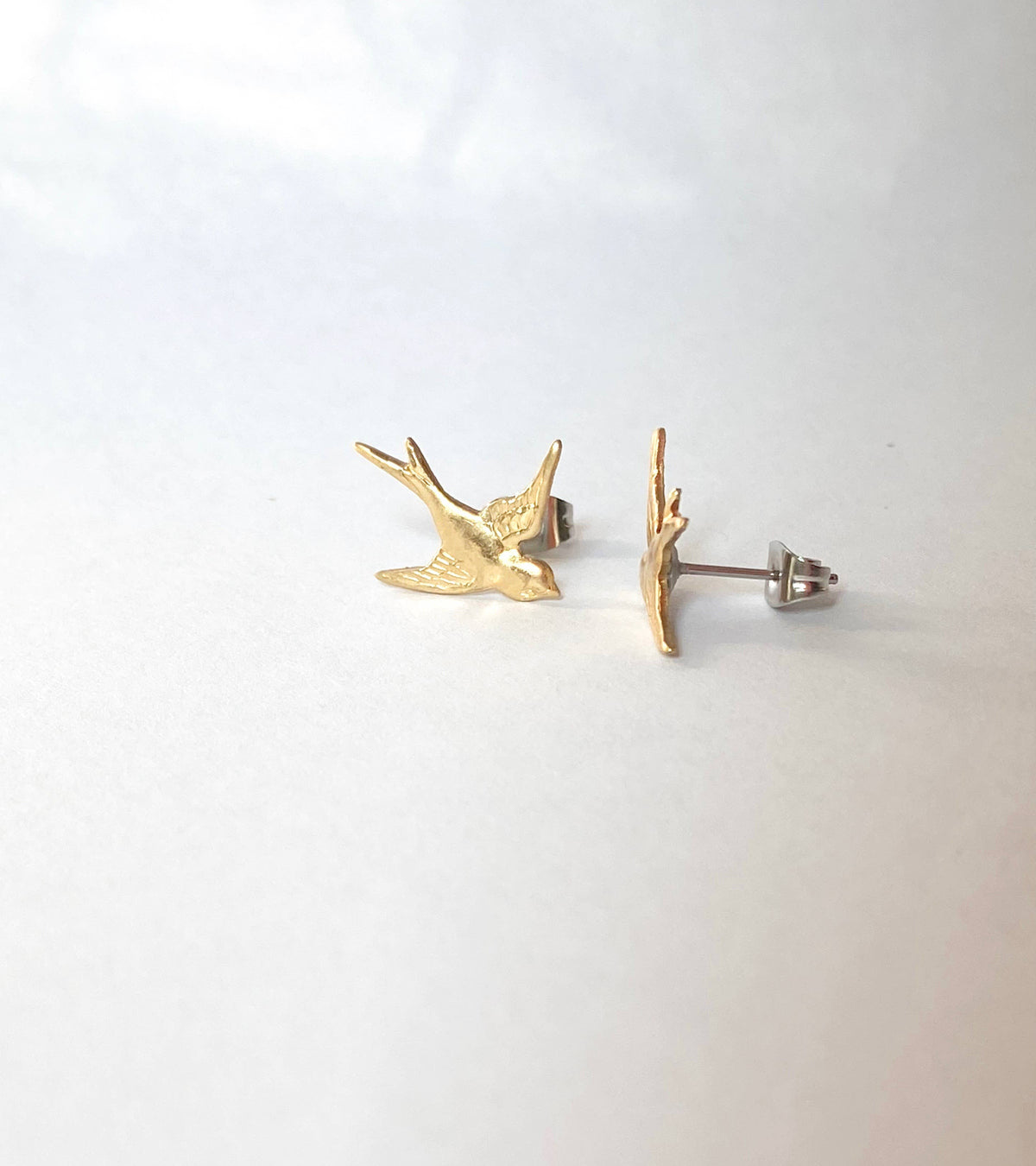 A pair of Flying Sparrow Earrings, crafted from golden brass, are displayed on a white background—one earring lies flat while the other stands on its edge. These nature-inspired accessories make a charming addition to your look, perfect for adding a whimsical touch to your woodland wedding ensemble.