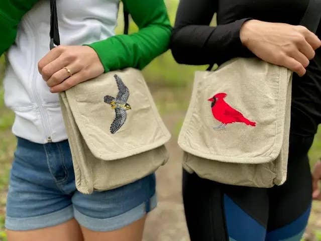 Two people stand outdoors, each with a handmade cotton Luna Moth Field Bag featuring an embroidered bird. The left bag shows a gray bird in flight; the right displays a bright red cardinal. Both individuals are dressed casually.