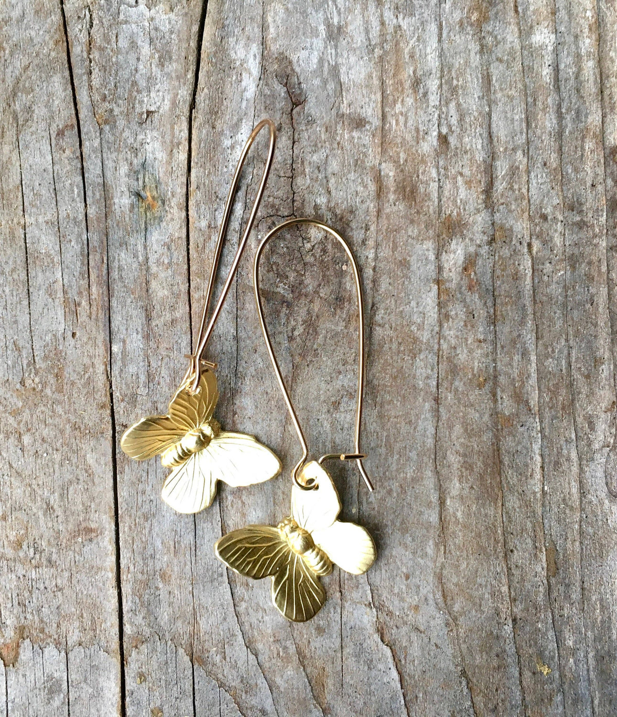 Two gold SmallButterfly Earrings with long hooks rest on a weathered wooden surface, showcasing delicate butterfly wing details against a rustic background. These nature-inspired earrings beautifully capture the elegance of butterfly charms, making them a perfect accessory for any occasion.