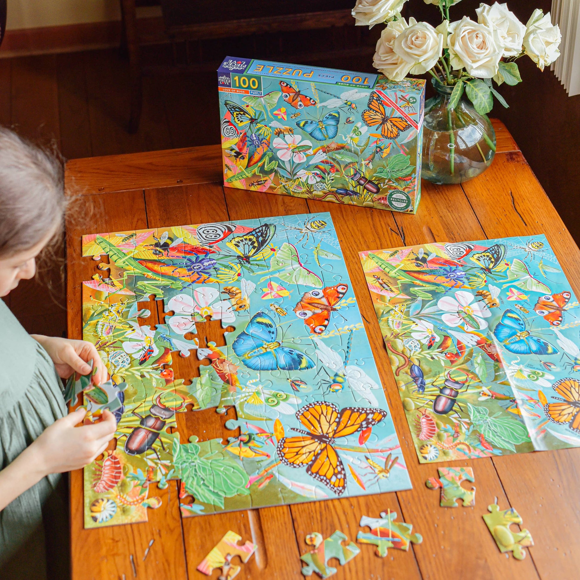 A child assembles the Love of Bugs 100 Piece Puzzle, featuring butterflies and bugs, on a wooden table. The completed puzzle, its box, and a vase of white roses sit nearby as bright natural light fills the room.