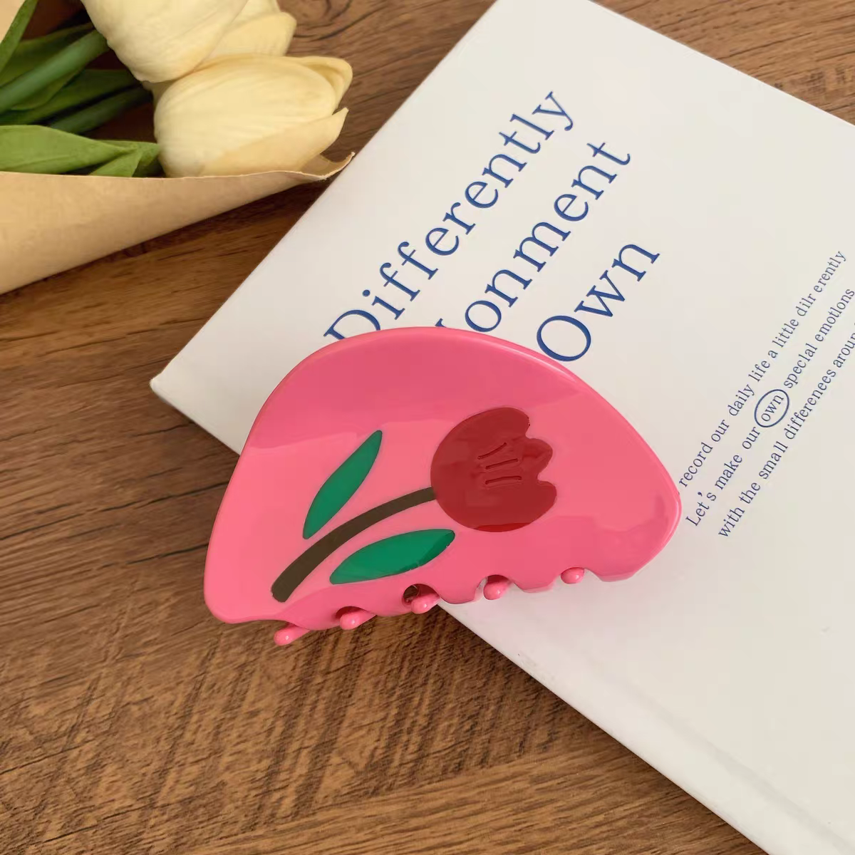 A pink, semi-circular Tulip Hair Claw Clip decorated with a red tulip and green leaves, resting on a white book.