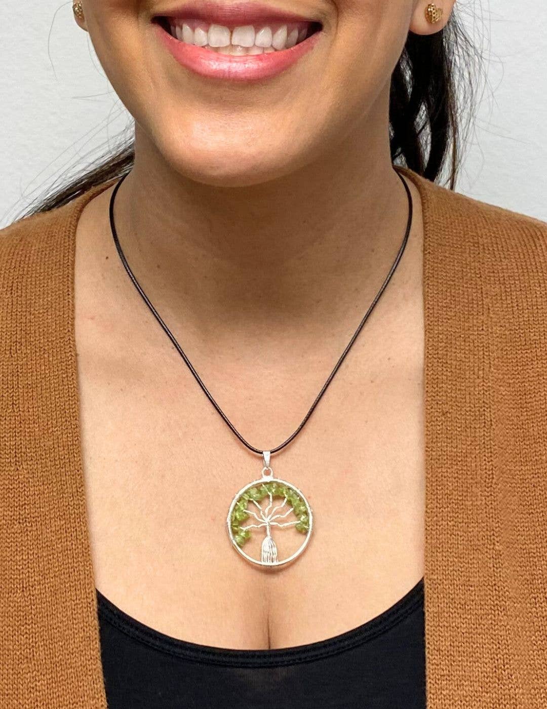 A woman, smiling and wearing small round earrings, is dressed in a brown cardigan and a black cord necklace featuring the Silver Plated Tree of Life Stone Pendant adorned with green stones.