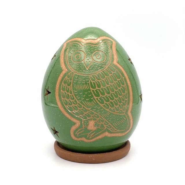 The Owl Luminary is a green, egg-shaped ceramic piece with a gold owl design and star cutouts, set on a small brown circular base against a white background, highlighting exquisite handcrafted detail.