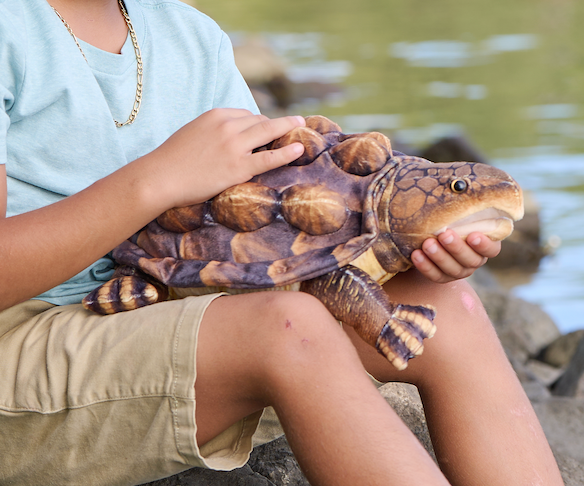 A child sitting outdoors by water holds the Wild Onez Snapping Turtle 15″ (38cm) stuffed animal on their lap, making it a perfect addition for anyone seeking unique swamp-themed nursery decor.