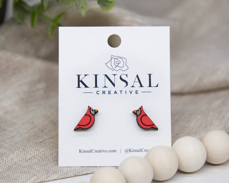 Cardinal Wood Stud Earrings in the shape of red cardinals are displayed on a white Kinsal Creative card, with wooden beads and green leaves in the background—a charming handmade jewelry choice.