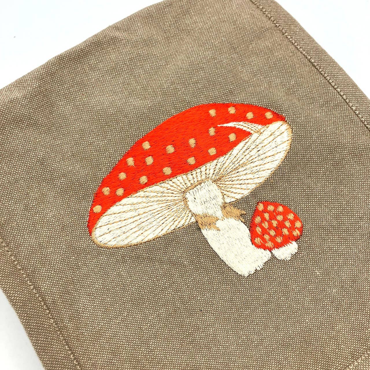 The Fly Agaric Mushroom Field Bag features an embroidered image of two red-capped, white-stemmed mushrooms—one large, one small—each with yellowish-white spots on a light brown handmade cotton bag.