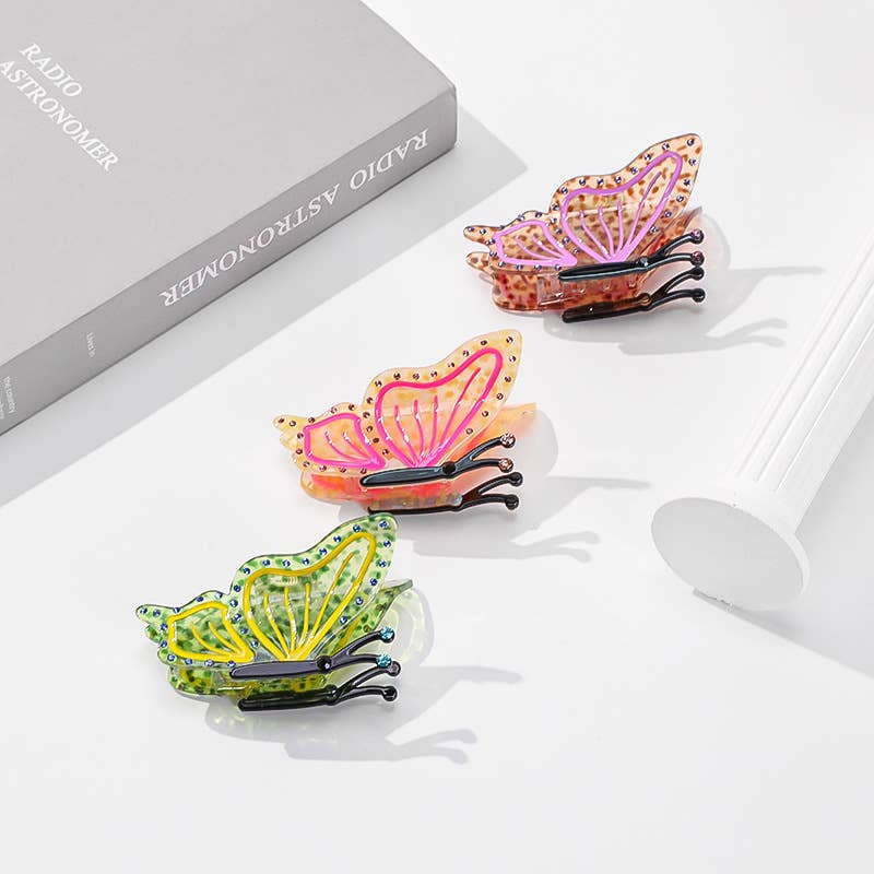 Three pink, orange, and green Butterfly Hair Claw clips with black details resting on a white surface next to a book.