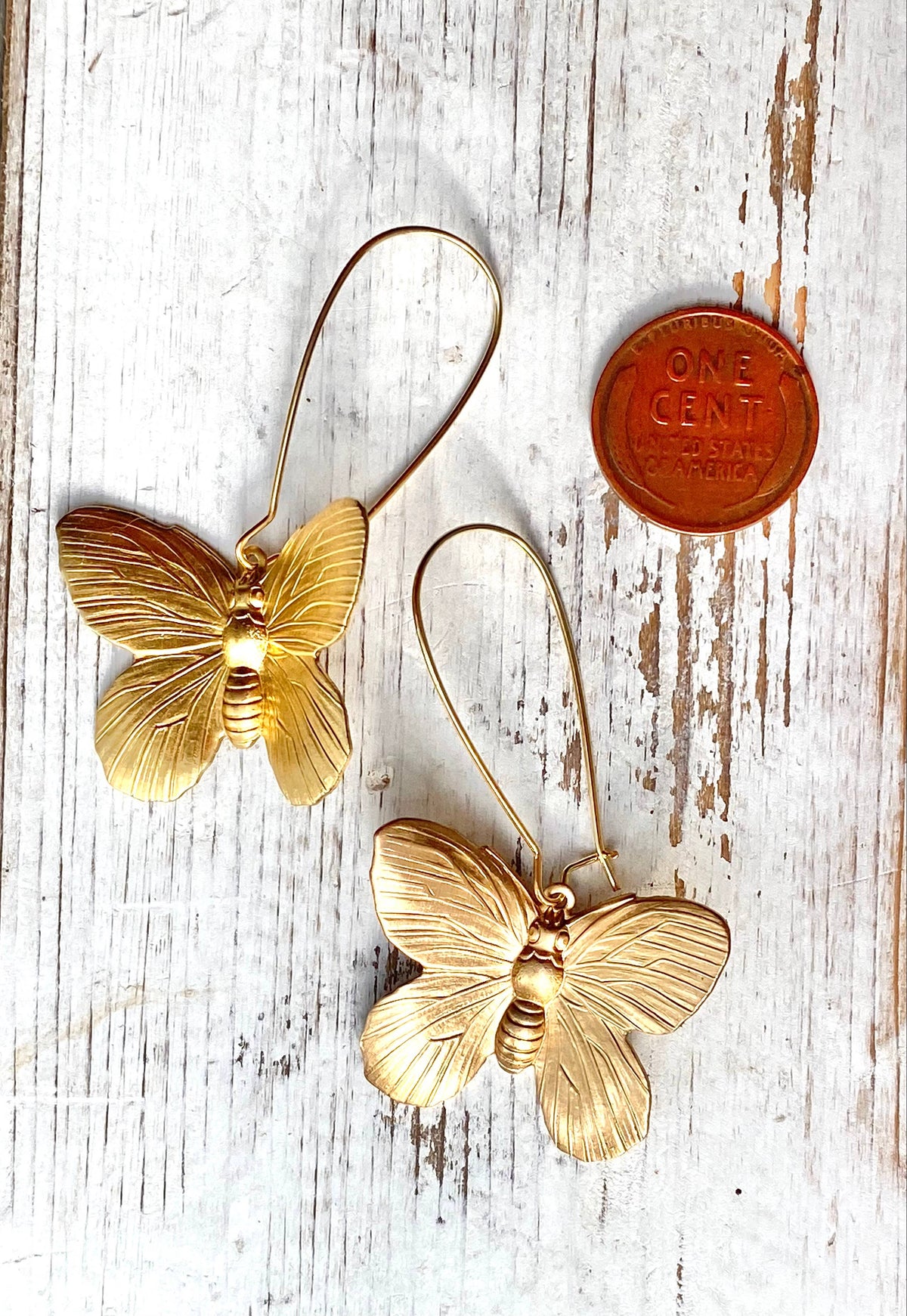 Two Butterfly Earrings are displayed on a rustic white wooden surface next to a U.S. one cent coin for size comparison, highlighting the delicate statement design and charming butterfly motif.
