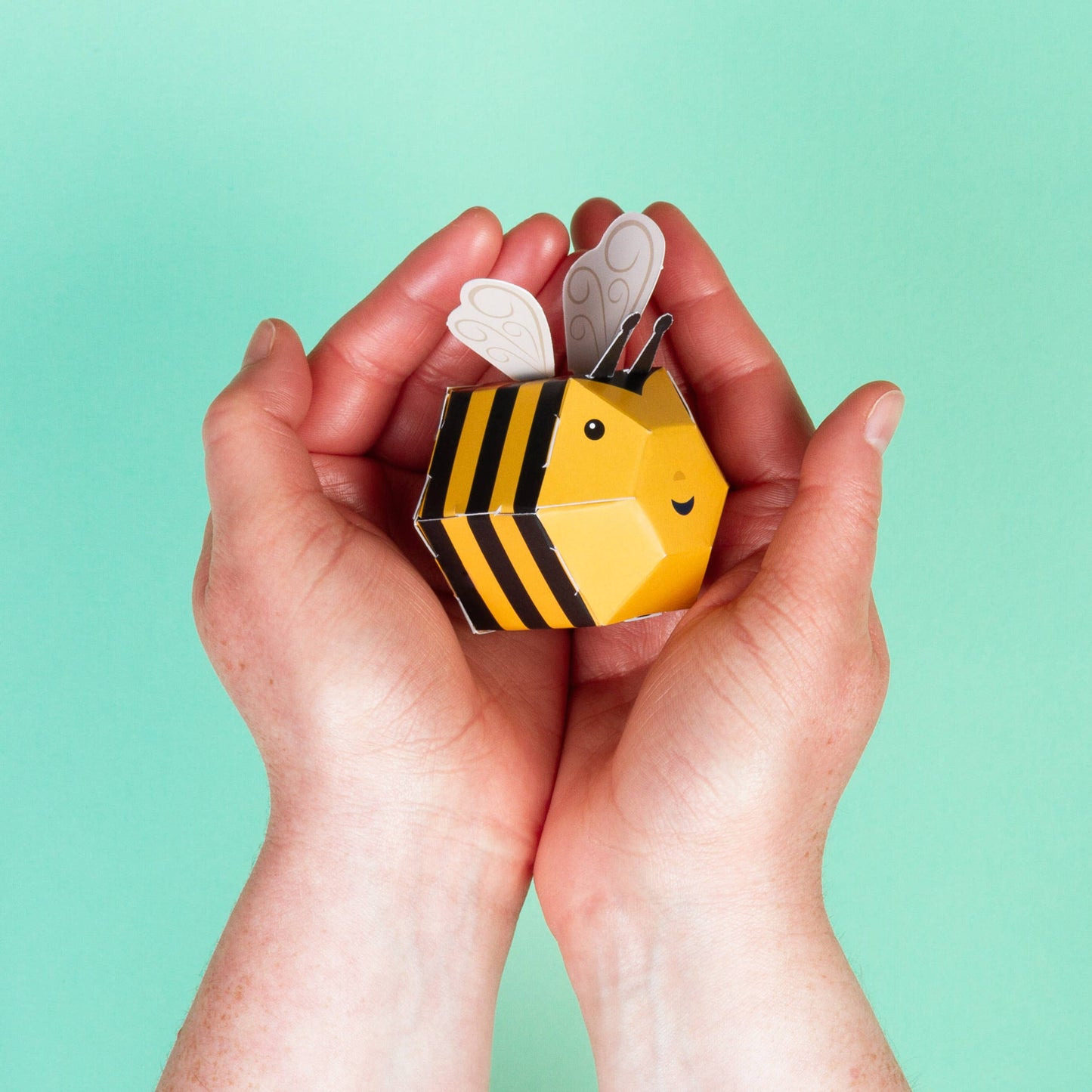 A pair of hands gently cups a Create Your Own Buzzy Bumble Bee—featuring black stripes, a smiling face, and gray wings—against a turquoise background. This plastic-free kit is ideal for creative fun.