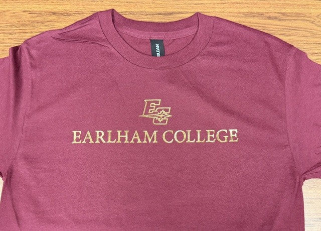 The Gold Foil Tee features "EARLHAM COLLEGE" and a stylized E with a star, printed in gold on the chest of a maroon T-shirt, shown laid flat on a wooden surface.