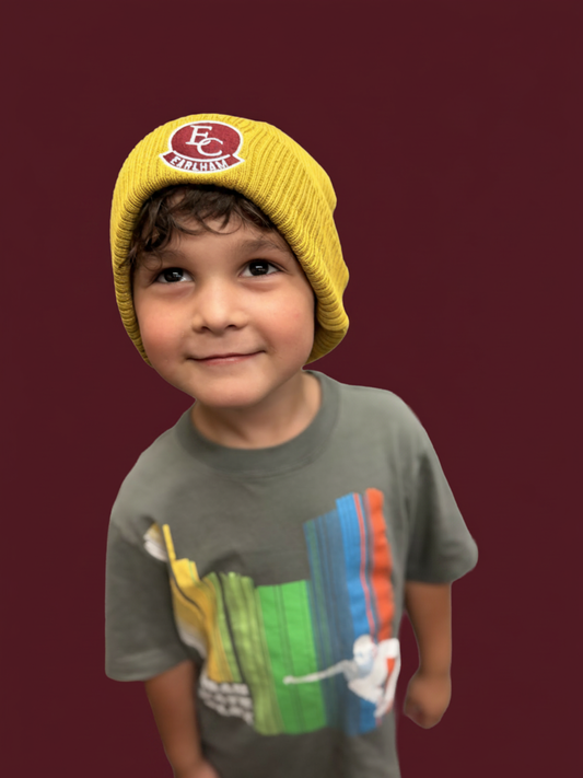 A young child smiles and looks up while wearing the Golden Rollup Beanie—a yellow hat featuring a maroon and white patch—paired with a gray t-shirt with a colorful graphic, set against a solid maroon background.