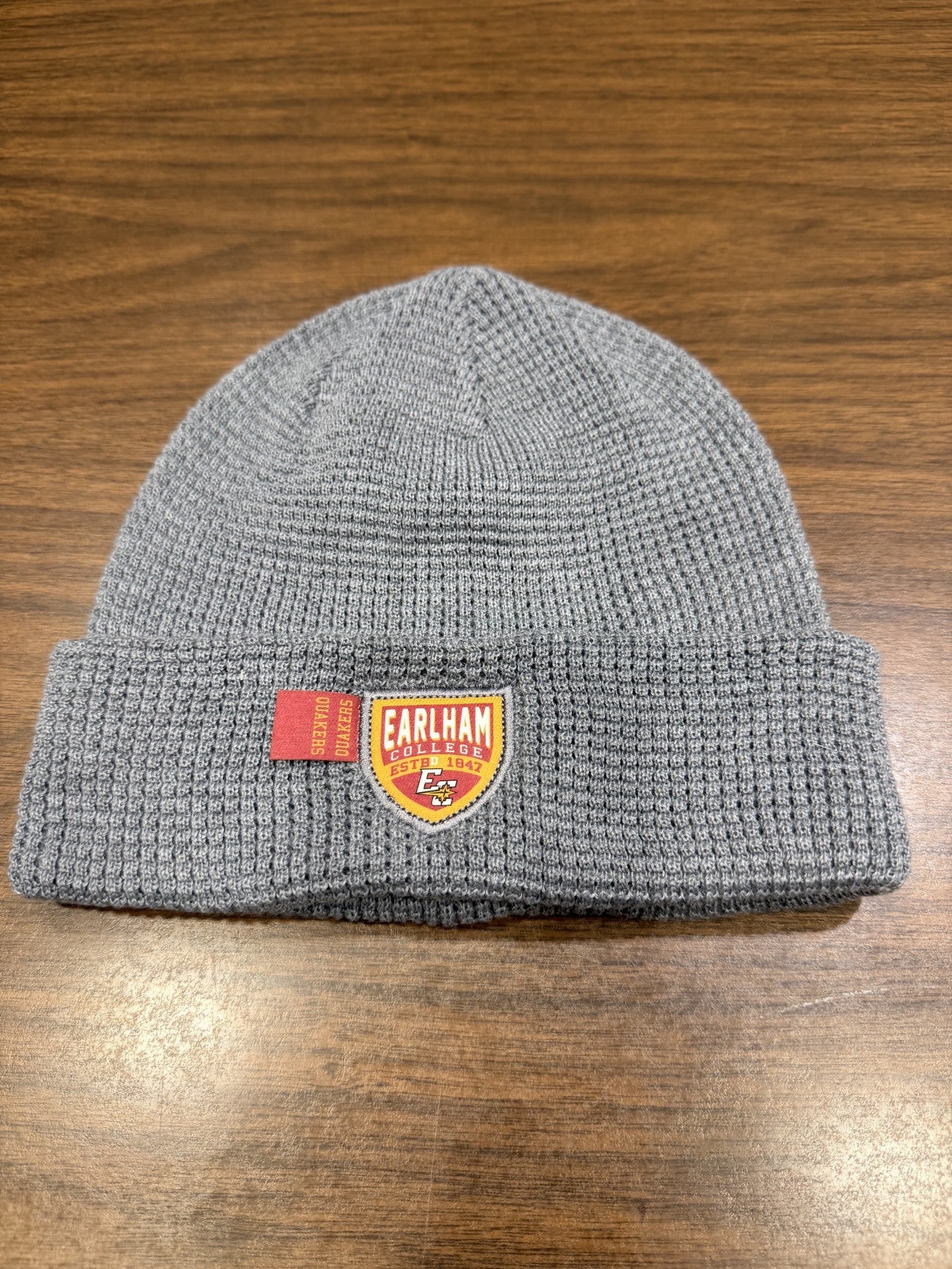 A Waffle Knit Cuff Beanie in gray featuring an Earlham College patch and a small red tag, displayed on a wooden surface.