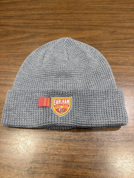 A Waffle Knit Cuff Beanie in gray featuring an Earlham College patch and a small red tag, displayed on a wooden surface.