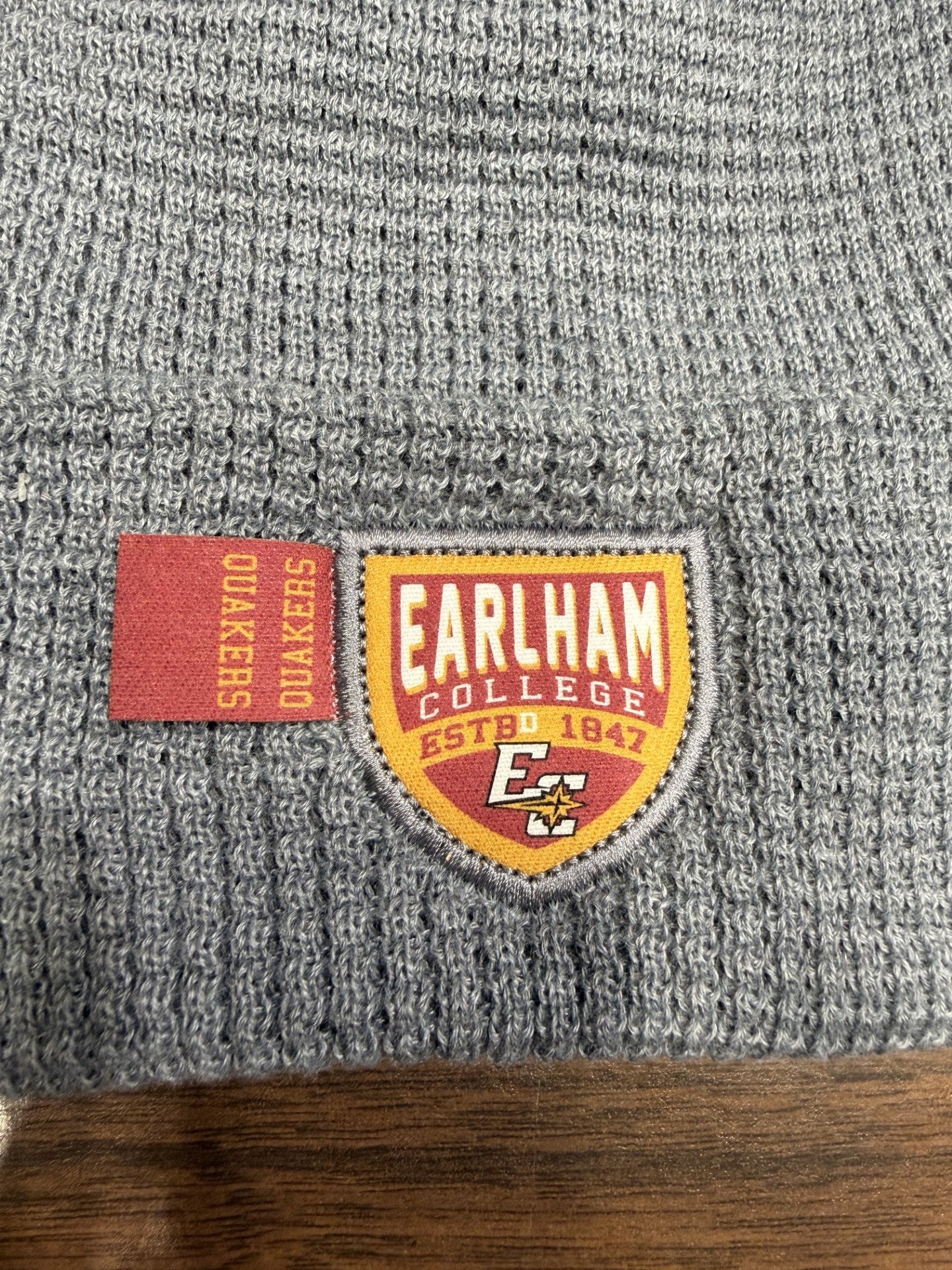 A close-up of the Waffle Knit Cuff Beanie in gray, featuring a sewn badge with "Earlham College Estbd 1847" and "Quakers" on a red tag, along with the school logo and colors.