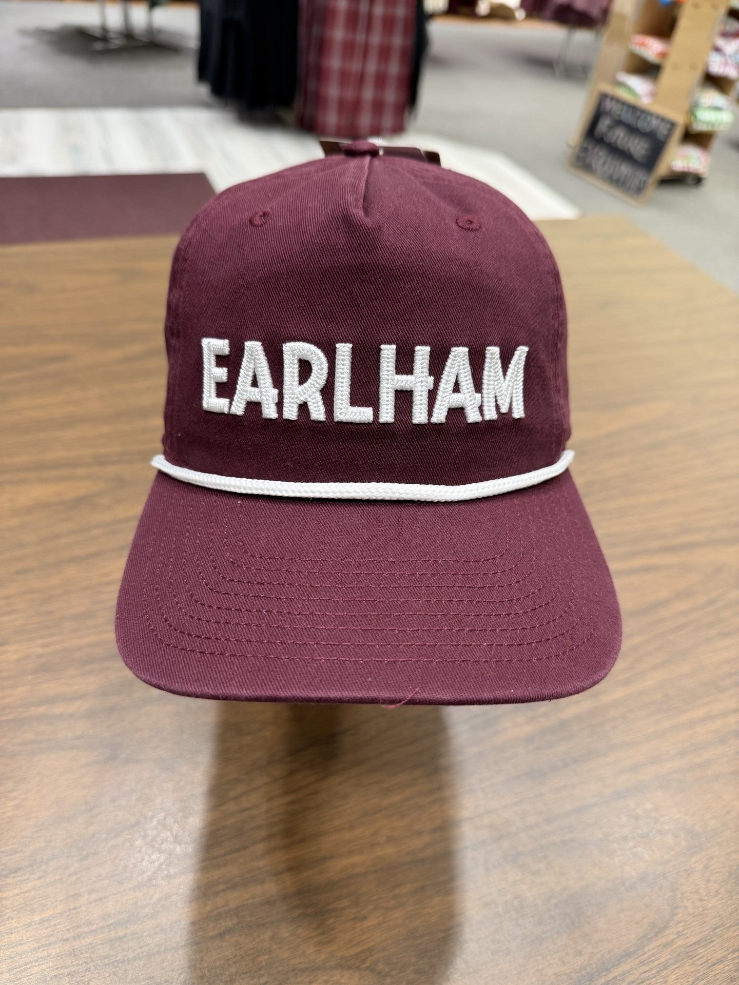 An Earlham Rope Hat in maroon, featuring “EARLHAM” embroidered in large white letters on the front, is displayed on a wooden table indoors.