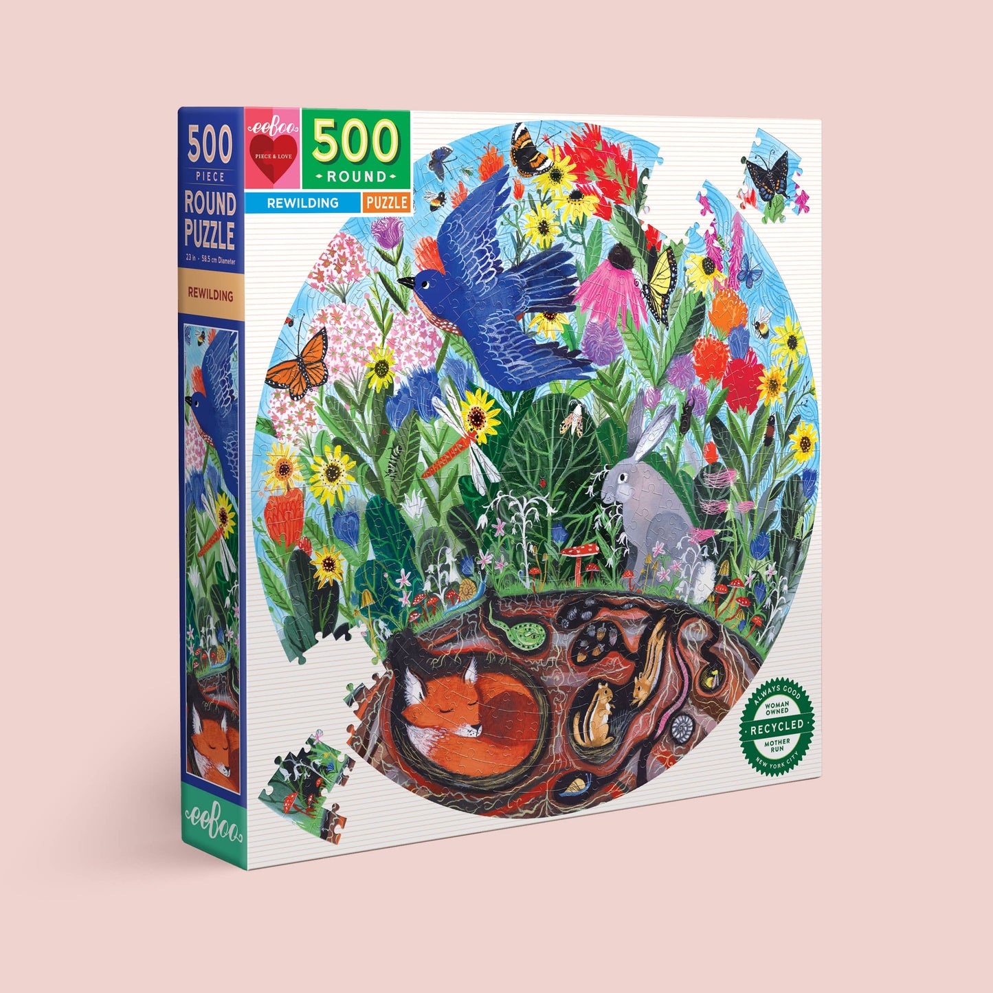 The Rewilding 500 Piece Puzzle comes in a round box with a vibrant garden scene—meadow flowers, bluebird, butterflies, rabbit, and a fox in its burrow. The box is labeled Rewilding and features a recycled materials seal.