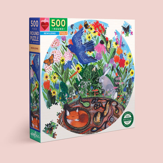 The Rewilding 500 Piece Puzzle comes in a round box with a vibrant garden scene—meadow flowers, bluebird, butterflies, rabbit, and a fox in its burrow. The box is labeled Rewilding and features a recycled materials seal.